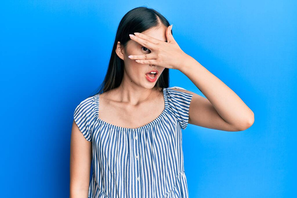 Jonge Chinese vrouw draagt casual gestreept t-shirt gluren in shock bedekking gezicht en ogen met de hand, kijken door vingers met beschaamde uitdrukking.  - Foto, afbeelding