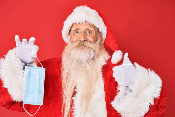 Παλιά ανώτερος άνθρωπος φορώντας κοστούμι Santa Claus κρατώντας μάσκα ασφαλείας χαμογελώντας χαρούμενος και θετικός, αντίχειρας επάνω κάνει εξαιρετική και την έγκριση σημάδι  - Φωτογραφία, εικόνα