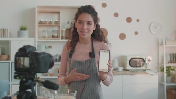 Die junge kaukasische Video-Bloggerin in Schürze steht in der Küche, spricht, lächelt und zeigt ein Smartphone mit weißem Bildschirm auf einer semiprofessionellen Kamera, die vor ihr steht - Filmmaterial, Video