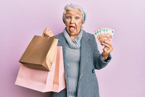 Женщина с седыми волосами, держащая сумки с покупками и банкноты евро, торчащие языком, довольна смешным выражением лица..  - Фото, изображение