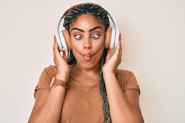 Joven mujer afroamericana con trenzas escuchando música usando auriculares haciendo cara de pez con boca y ojos entrecerrados, loca y cómica.  - Foto, imagen