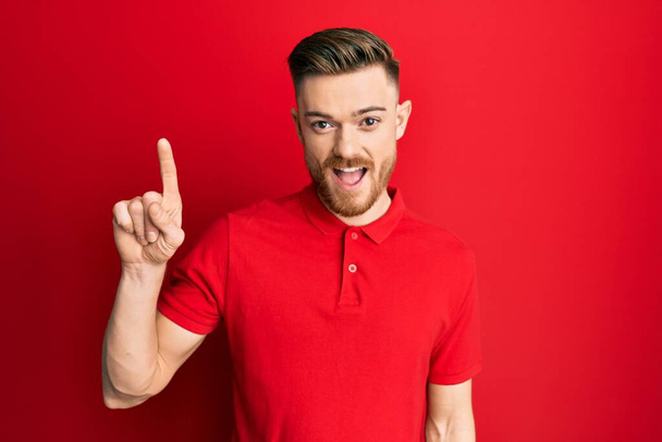 Fiatal vörös hajú férfi alkalmi ruhát visel mosolyog egy ötlettel vagy kérdéssel mutogatva ujjal felfelé boldog arccal, első számú  - Fotó, kép