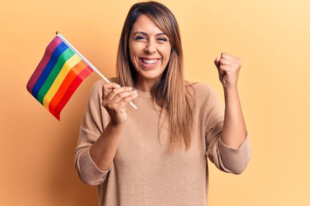 Young beautiful woman holding lgtbq flag screaming proud, celebrating victory and success very excited with raised arm  - Photo, Image