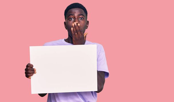 Junger afrikanisch-amerikanischer Mann mit leeren Transparenten, die den Mund mit der Hand bedecken, schockiert und ängstlich vor Irrtum. Überraschte Miene  - Foto, Bild
