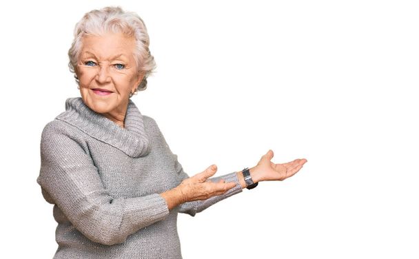 Ältere grauhaarige Frau in lässigem Winterpullover lädt ein, lächelnd natürlich mit offener Hand einzutreten  - Foto, Bild