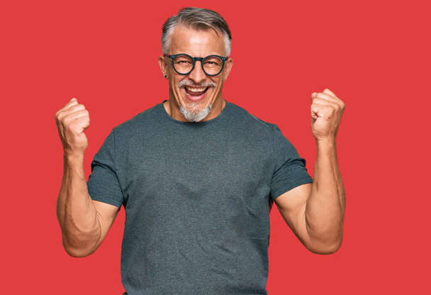 Grauhaariger Mann mittleren Alters in lässiger Kleidung und Brille schreit stolz und feiert Sieg und Erfolg mit erhobenen Armen  - Foto, Bild