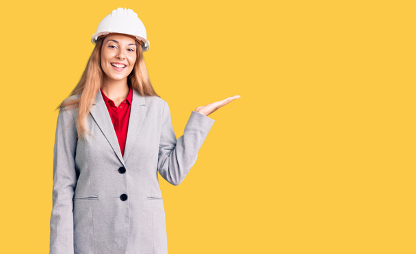 Beautiful young woman wearing architect hardhat smiling cheerful presenting and pointing with palm of hand looking at the camera.  - Photo, Image