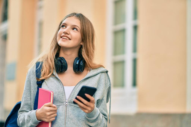 Schöne kaukasische Studentin lächelt glücklich mit Smartphone in die Stadt. - Foto, Bild