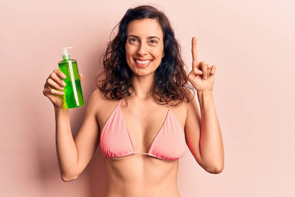 Joven mujer hispana hermosa usando bikini con loción de aloe vera sonriendo con una idea o pregunta apuntando con el dedo con la cara feliz, número uno  - Foto, imagen