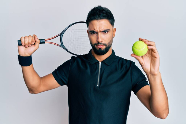 Молодой человек с бородой играет в теннис держа ракетку и мяч невежественное и запутанное выражение лица. Концепция сомнений.  - Фото, изображение