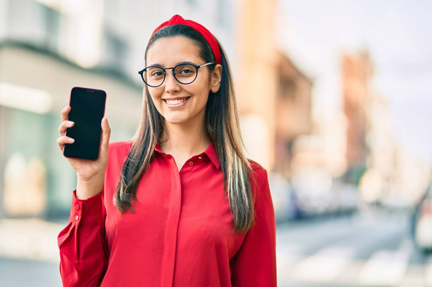 Fiatal spanyol nő mosolyog boldog bemutató képernyő okostelefon a városban. - Fotó, kép