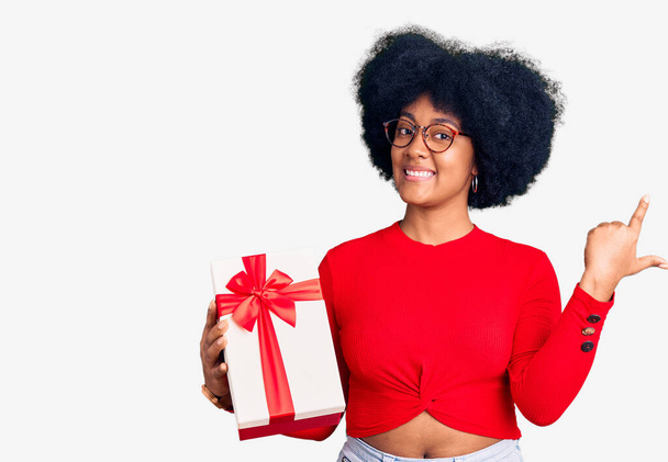Νεαρή Αφρικάνα Αμερικανίδα που κρατάει το δώρο της χαρούμενο με χαμόγελο στο πρόσωπο δείχνοντας με το χέρι και το δάχτυλο στο πλάι με χαρούμενη και φυσική έκφραση  - Φωτογραφία, εικόνα