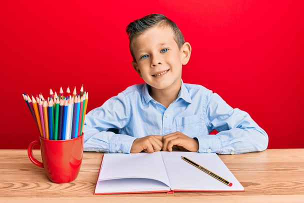 Uroczy biały dzieciak piszący książkę na stole ze szczęśliwym i chłodnym uśmiechem na twarzy. szczęśliwy człowiek.  - Zdjęcie, obraz