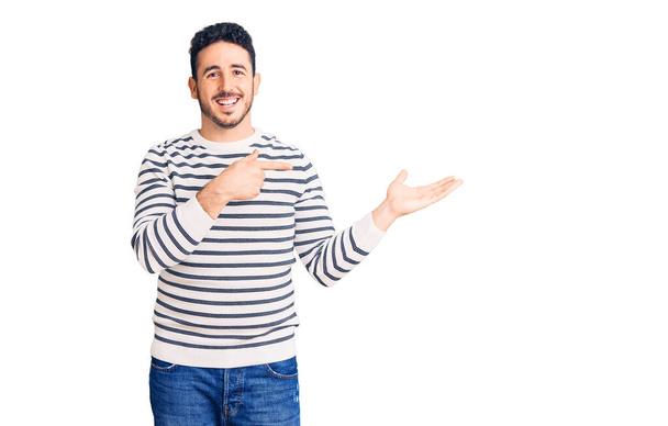 Νεαρός Ισπανός άνδρας φορώντας καθημερινά ρούχα κατάπληκτος και χαμογελώντας στην κάμερα ενώ παρουσιάζει με το χέρι και δείχνοντας με το δάχτυλο.  - Φωτογραφία, εικόνα