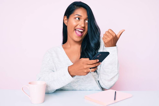 Красивая латинская молодая женщина с длинными волосами с помощью смартфона сидит на столе указывая большой палец вверх в сторону улыбается счастливо с открытым ртом  - Фото, изображение