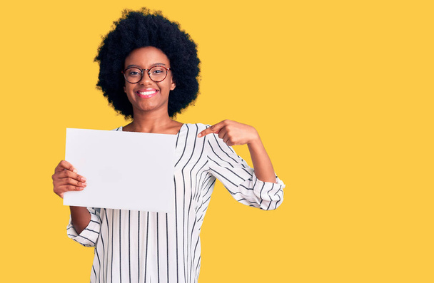 Молодая африканская американка держит пустую пустую бумагу, указывая пальцем на себя улыбающуюся счастливую и гордую  - Фото, изображение