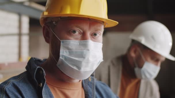 Retrato de cerca del arquitecto masculino en máscara facial protectora y sombrero de señora posando para la cámara mientras trabajaba en el sitio de construcción durante la pandemia de coronavirus - Imágenes, Vídeo
