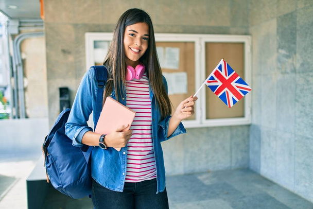Mladý hispánský student dívka usměvavý šťastný držení knihy a uk vlajka na univerzitě. - Fotografie, Obrázek
