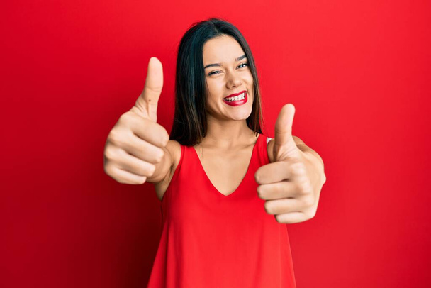 junges hispanisches Mädchen in lässigem Stil mit ärmellosem Hemd, das zustimmt, positive Geste mit der Hand zu machen, Daumen hoch lächelnd und glücklich über den Erfolg. Siegergeste.  - Foto, Bild