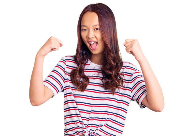 Młoda piękna chińska dziewczyna w luźnych ubraniach krzyczy dumny, świętując zwycięstwo i sukces bardzo podekscytowany z podniesionymi ramionami  - Zdjęcie, obraz