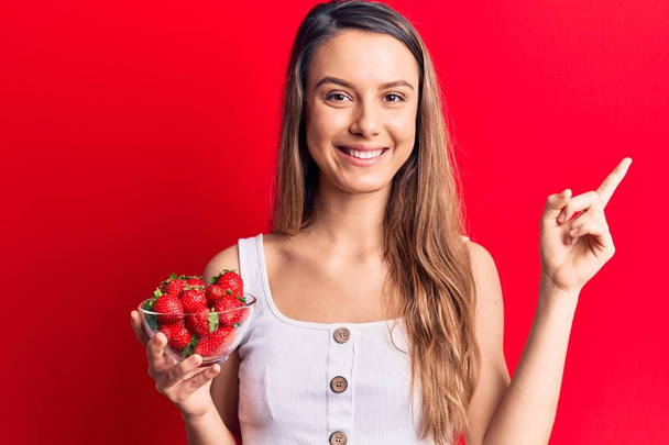 Jeune belle fille tenant bol avec des fraises souriant heureux pointant avec la main et le doigt sur le côté  - Photo, image