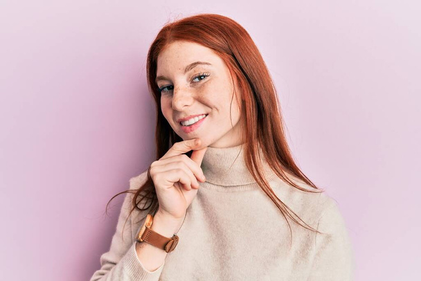 Young red head girl wearing turtleneck sweater smiling looking confident at the camera with crossed arms and hand on chin. thinking positive.  - Fotoğraf, Görsel