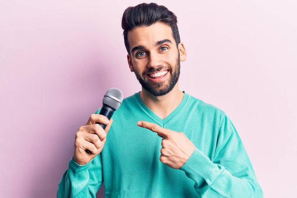 Молодий красивий чоловік з бородою співає пісню, використовуючи мікрофон усміхнений щасливий вказуючи рукою і пальцем
  - Фото, зображення