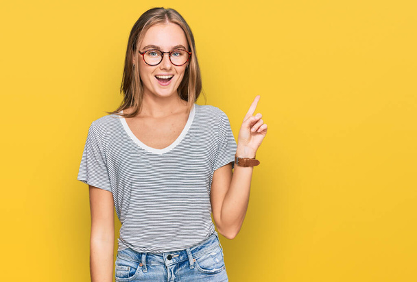 Schöne junge blonde Frau in lässiger Kleidung und Brille zeigt und zeigt mit Finger Nummer eins, während sie selbstbewusst und glücklich lächelt.  - Foto, Bild