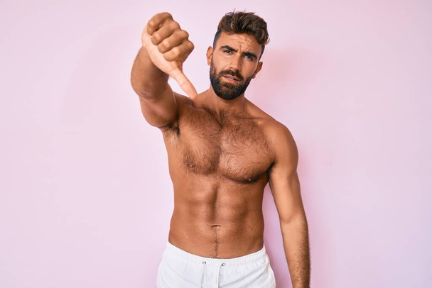 Young hispanic man standing shirtless looking unhappy and angry showing rejection and negative with thumbs down gesture. bad expression.  - Foto, imagen