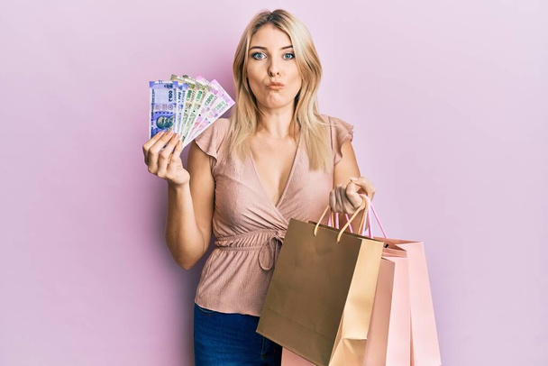 Young caucasian woman holding indian rupee banknotes and shopping bags making fish face with mouth and squinting eyes, crazy and comical.  - Foto, imagen