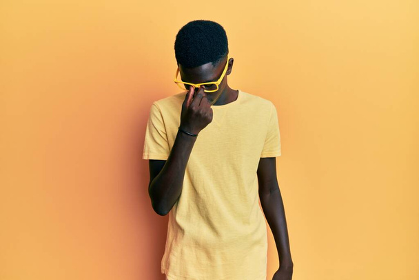 Молодой африканский американец в повседневной одежде и очках устал тереть нос и глаза, чувствуя усталость и головную боль. концепция стресса и разочарования.  - Фото, изображение