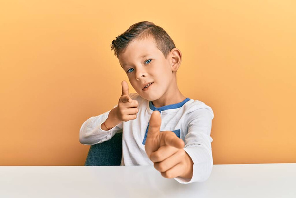 Roztomilé bělošské dítě, oblečené v neformálním oblečení, sedí na stole a ukazuje prsty na kameru se šťastnou a zábavnou tváří. dobrá energie a vibrace.  - Fotografie, Obrázek