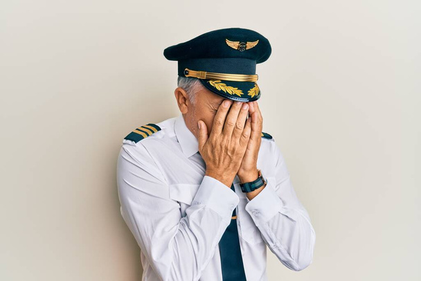 Schöner Mann mittleren Alters in Flugzeugpilotenuniform mit traurigem Gesichtsausdruck, der weinend das Gesicht mit den Händen bedeckt. Depressionskonzept.  - Foto, Bild