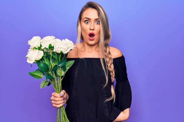Jonge mooie blonde vrouw met boeket bloemen over geïsoleerde paarse achtergrond bang en verbaasd met open mond voor verrassing, ongeloof gezicht - Foto, afbeelding