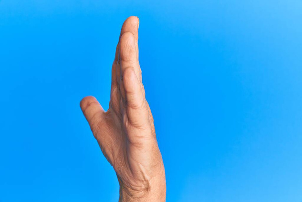 Mano del hombre hispano mayor sobre fondo azul aislado mostrando el lado de la mano estirada, empujando y haciendo gesto de parada  - Foto, Imagen