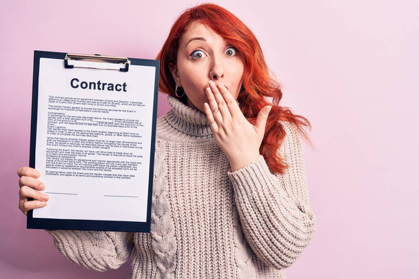 Young redhead woman holding clipboard with contract document paper over pink background covering mouth with hand, shocked and afraid for mistake. Surprised expression - Zdjęcie, obraz
