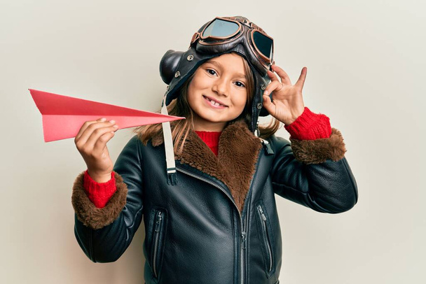 Little beautiful girl wearing pilot uniform holding paper plane doing ok sign with fingers, smiling friendly gesturing excellent symbol  - Foto, immagini