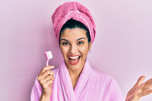 Νεαρή Ισπανίδα που φοράει μπουρνούζι και κρατάει οδοντόβουρτσα γιορτάζοντας το επίτευγμα με χαρούμενο χαμόγελο και έκφραση νικητή με σηκωμένο χέρι  - Φωτογραφία, εικόνα