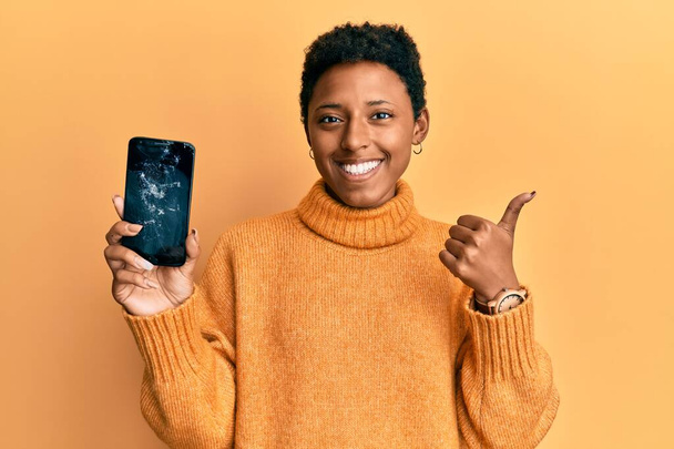 Молодая африканская американская девушка держит сломанный смартфон показывая треснувший экран улыбаясь счастливый и позитивный, большой палец вверх делает отлично и знак одобрения  - Фото, изображение