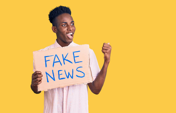Mladý africký Američan drží falešné zprávy prapor ukazuje palcem nahoru na stranu usmívá šťastný s otevřenými ústy  - Fotografie, Obrázek