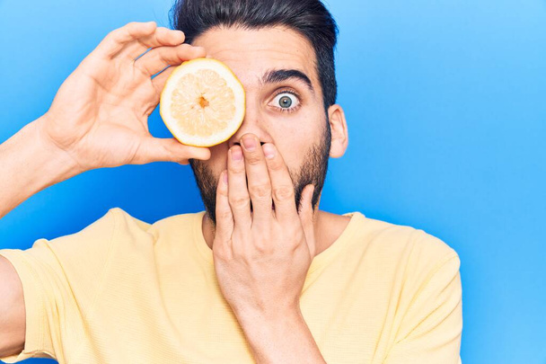 Mladý pohledný muž s plnovousem a s plátkem citronu přes oko zakrývajícím ústa rukou, šokovaný a vystrašený z omylu. překvapený výraz  - Fotografie, Obrázek