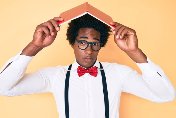 Bello uomo nerd afro-americano con i capelli afro tenendo libro sopra la testa espressione indiscreta e confusa. concetto di dubbio.  - Foto, immagini