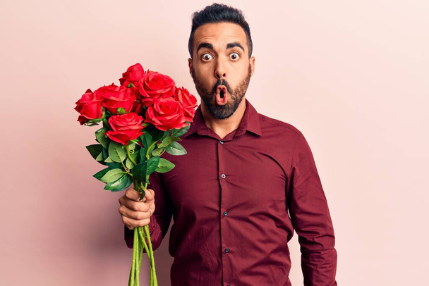 Jonge Spaanse man houdt bloemen bang en verbaasd met open mond voor verrassing, ongeloof gezicht  - Foto, afbeelding