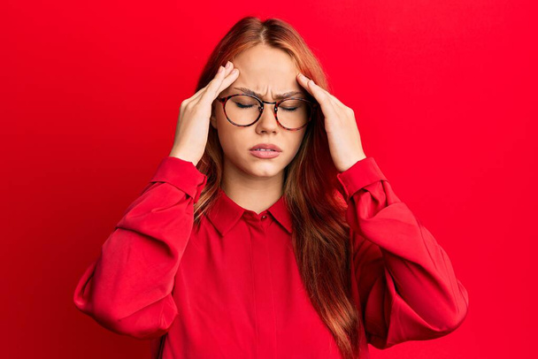 Nuori kaunis punapää nainen päällään rento vaatteita ja lasit yli punainen tausta käsi pään päälle kipua päähän, koska stressi. migreeni.  - Valokuva, kuva