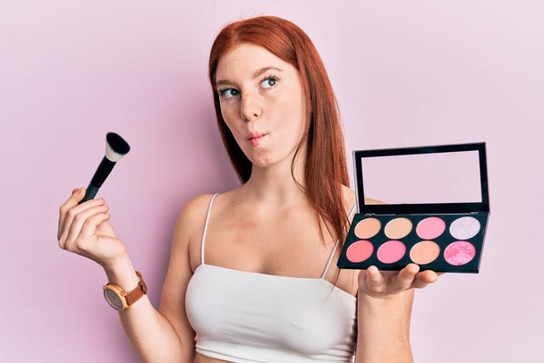 Young red head girl holding makeup brush and blush palette making fish face with mouth and squinting eyes, crazy and comical.  - Photo, Image