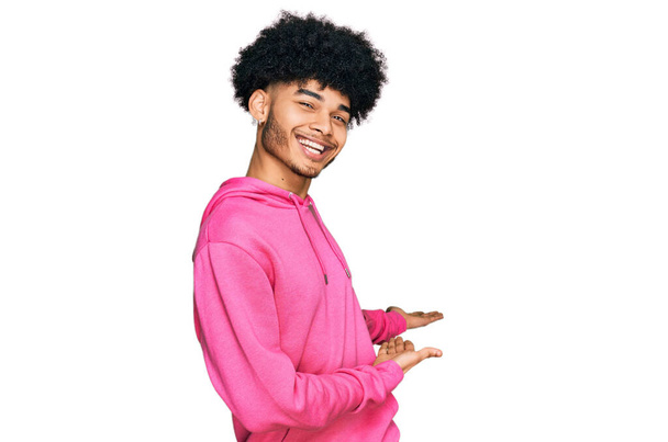 Молодой африканский американец с афроволосами в розовой толстовке приглашает войти улыбающимся натурально с открытой рукой  - Фото, изображение
