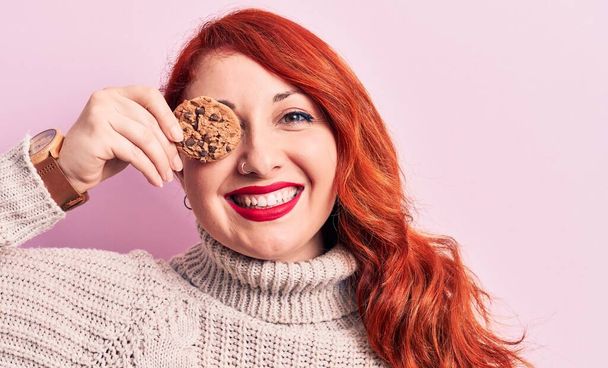 Young beautiful redhead woman standing holding chocolate cookie over eye looking positive and happy standing and smiling with a confident smile showing teeth - Zdjęcie, obraz