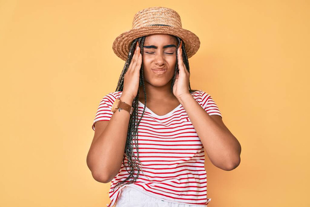 Young african american woman with braids wearing summer hat suffering from headache desperate and stressed because pain and migraine. hands on head.  - Foto, afbeelding