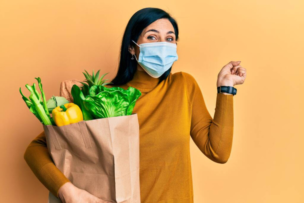 Young caucasian woman wearing medical mask holding groceries paper bag screaming proud, celebrating victory and success very excited with raised arm  - Photo, Image
