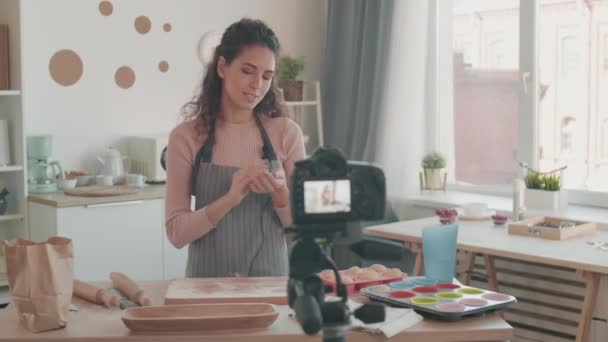 Средняя слежка слева от улыбающейся молодой кавказской женщины, стоящей у стола с деревянной посудой, простыней, полотенцем, делающей тестовые шарики, помещающей их в силиконовую форму, показывающей на своей камере - Кадры, видео